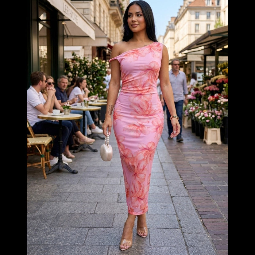 A woman wearing a pink floral one-shoulder maxi dress walking down a European-style street with outdoor cafes, representing the muse era collection from SLAY Boutique