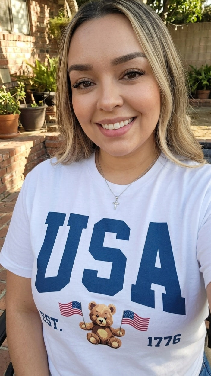 Woman wearing a white t-shirt with 'USA' and a teddy bear graphic outdoors.