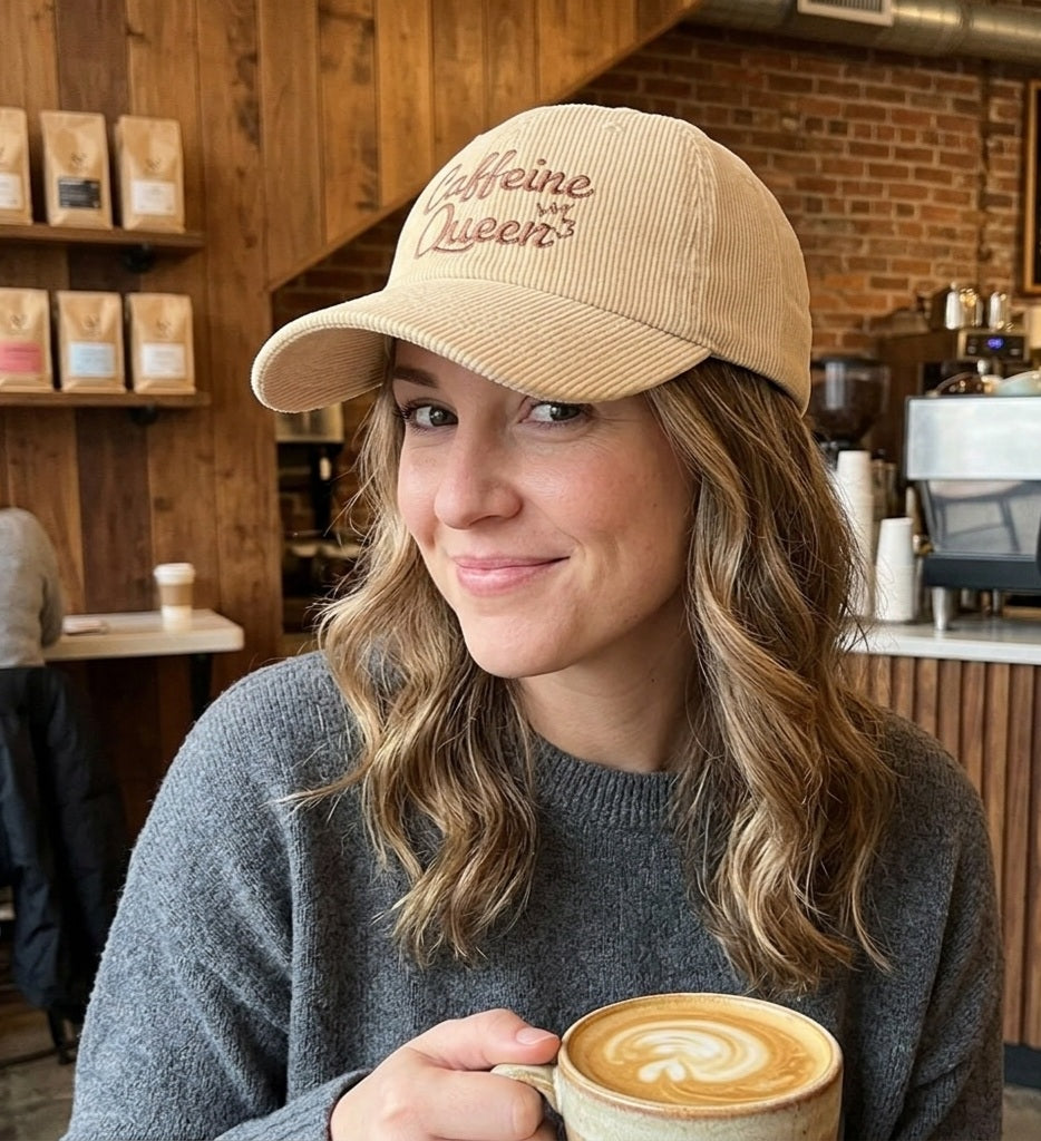 Woman wearing a 'Caffeine Queen' cap in a coffee shop holding a cup of coffee.