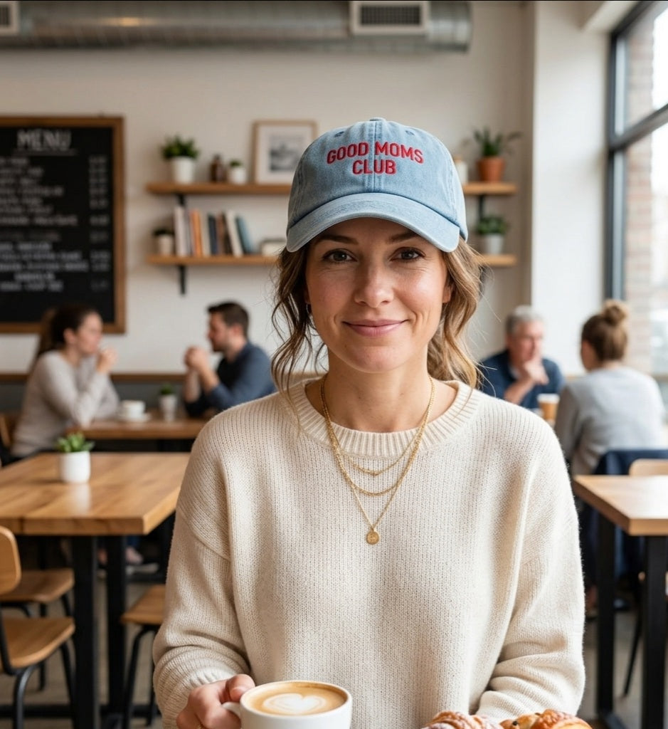 Woman wearing a 'Good Moms Club' cap in a coffee shop holding a cup.