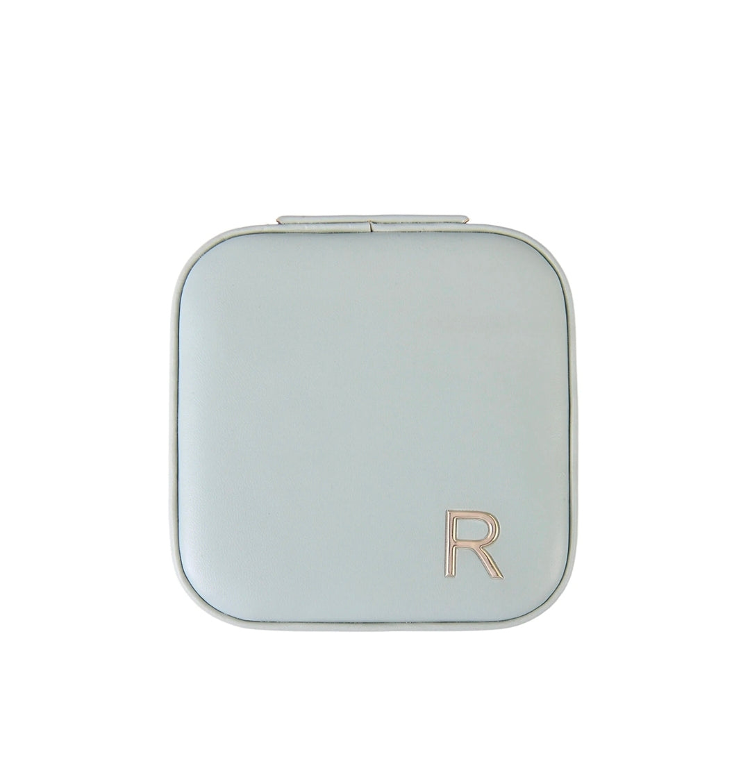 A square compact case with rounded corners in a pale sage green color, featuring a metallic rose gold letter 'R' logo in the bottom right corner, set against a plain white background.
