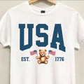 White t-shirt with 'USA' text and teddy bear graphic on a hanger.