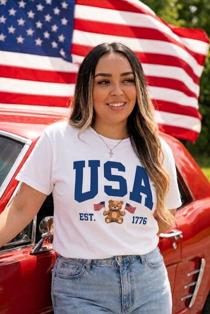 Woman wearing a 'USA' t-shirt with a teddy bear graphic, standing in front of an American flag and red car.